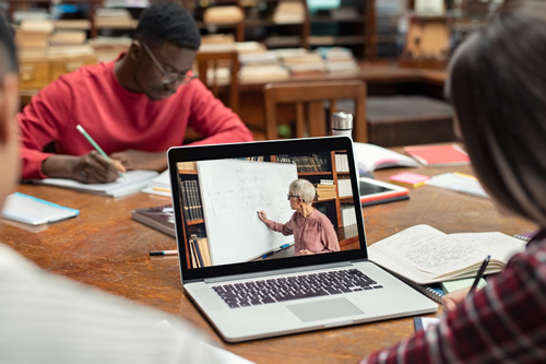 Two students sitting at a table taking notes and watching a lecture on a laptop