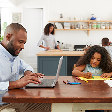 a family sitting in a kitchen with the father on a laptop, a daughter coloring at the table, and the mother and son standing near the counter table