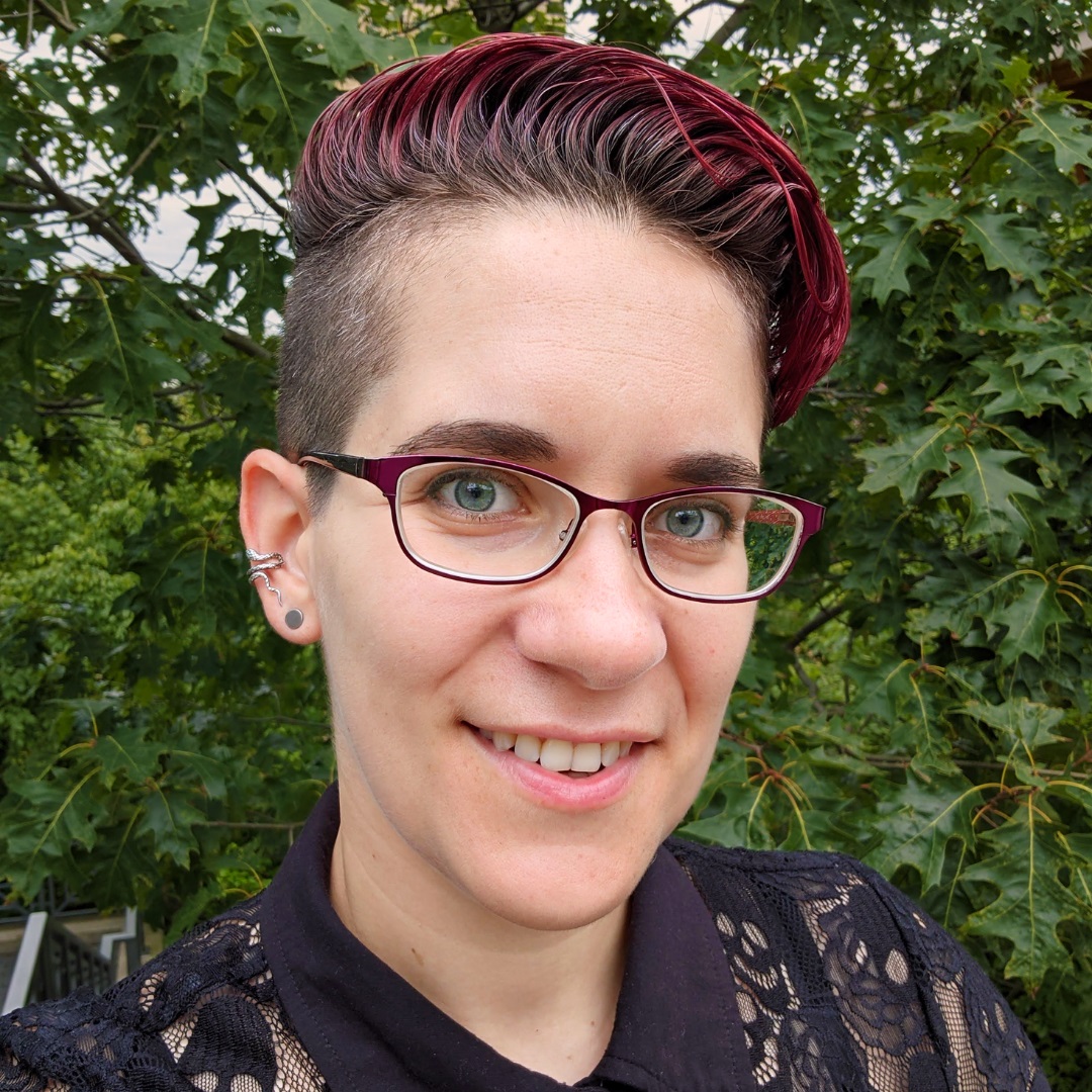 A photo of a white person with short hair buzzed on the sides. They wear a black button-front shirt, stud earrings, and a snake climber.